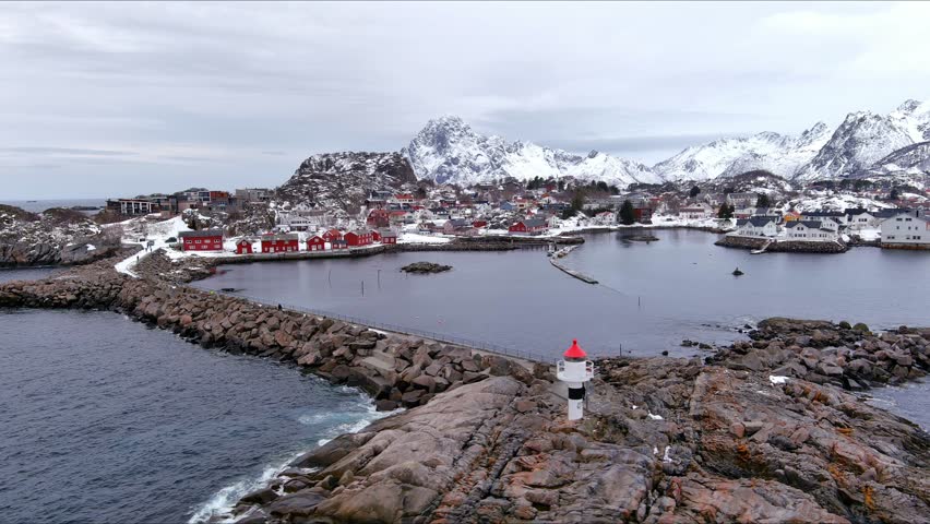 Aerial view of a small lighthouse on a rocky outcrop overlooking a coastal village with red cabins and snow-capped mountains in Nordland, Norway.