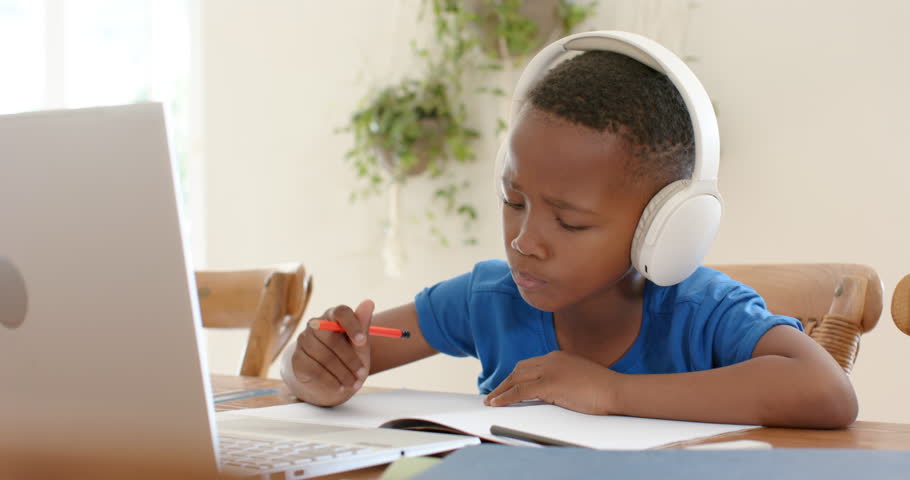 African American child writing notebook with pencil at home with headset raising hand to laptop cue. Student, headphones, learning, remote, education, focus, natural