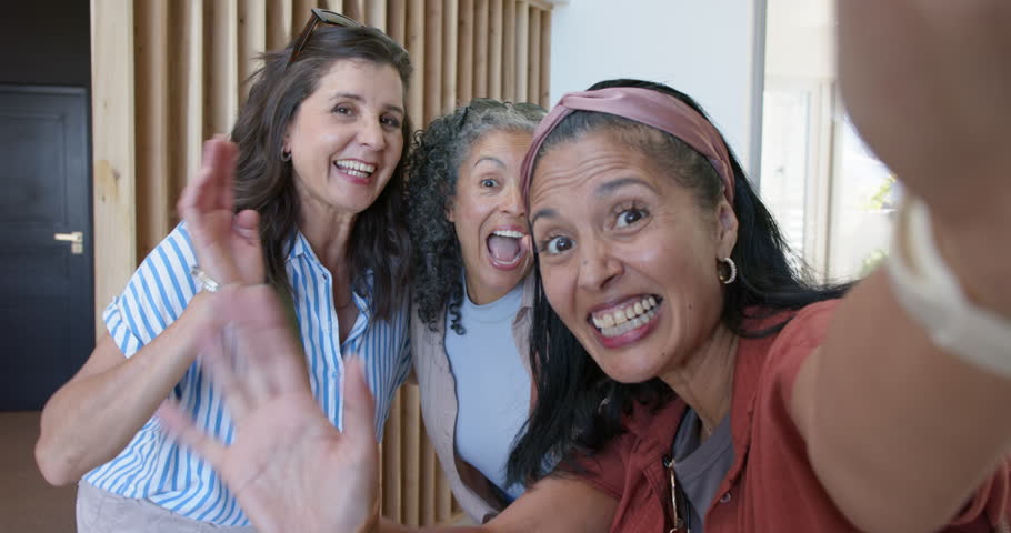 Diverse female friends leaning in as right woman holding smartphone recording selfie in home foyer. Trio, candid, laughter, entryway, natural, light, interior