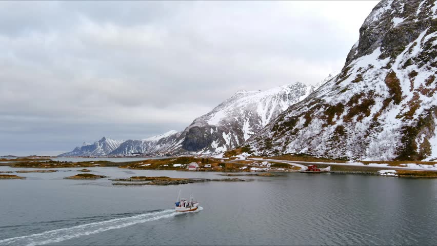 Aerial view of a fishing boat cruising through a fjord with snow-covered mountains and rocky islands under a cloudy sky in Nordland, Norway.