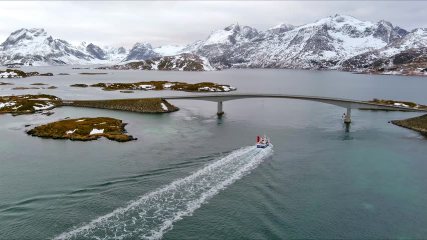 Aerial view of the Fredvang Bridges as a fishing boat leaves a wake in the water surrounded by snow-capped mountains in Nordland, Norway.