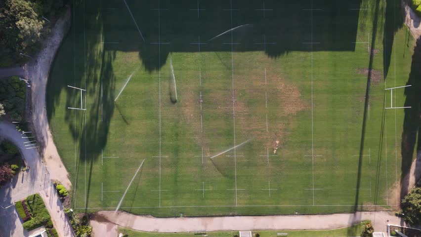 Aerial view of a green rugby pitch with active sprinklers watering the grass and white line markings during a sunny day.