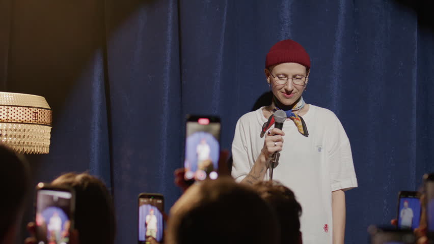 Young female comedian holding microphone and performing stand up set on stage as audience recording show with their smartphones