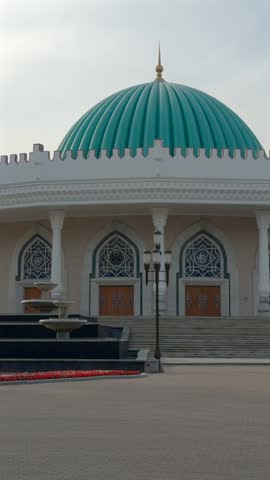 Front view of State Museum of Temurids History in Tashkent with turquoise dome, arched windows and columns, steps and fountain in foreground in soft daylight.