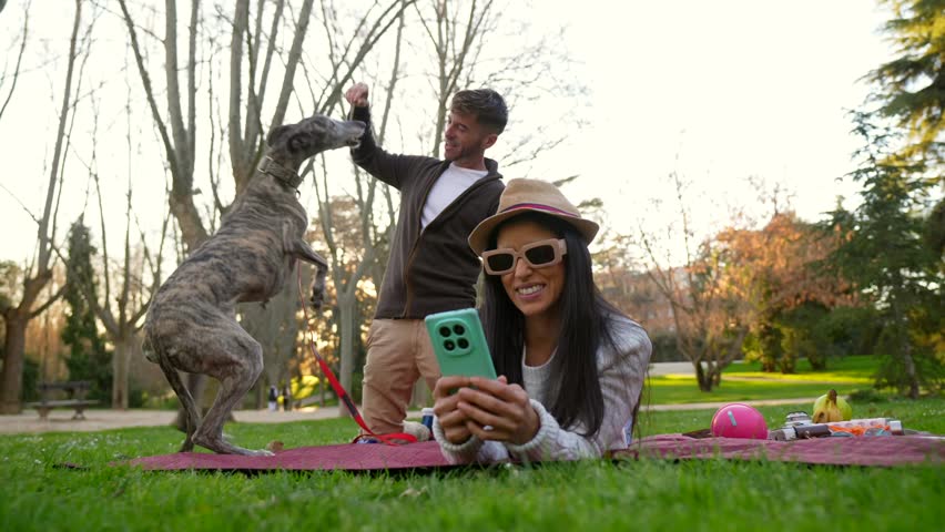 Happy woman wearing sunglasses and a hat taking a selfie with her phone while lying on a picnic blanket, as her boyfriend plays with their greyhound dog in the background on a sunny day