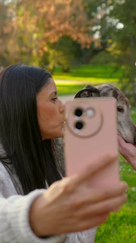 Happy young couple taking a selfie with their adorable greyhound dog outdoors in a sunny park, with the man kissing the pet while the woman holds the smartphone and smiles for the photo
