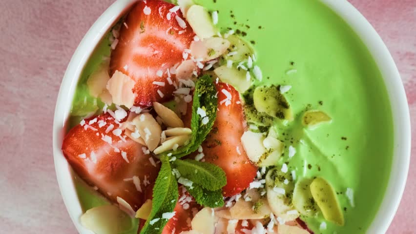 Green tea matcha yogurt topped with strawberries, mint and almond flakes on pastel pink background. Top view. Healthy dessert, protein rich smoothie breakfast. Flat lay