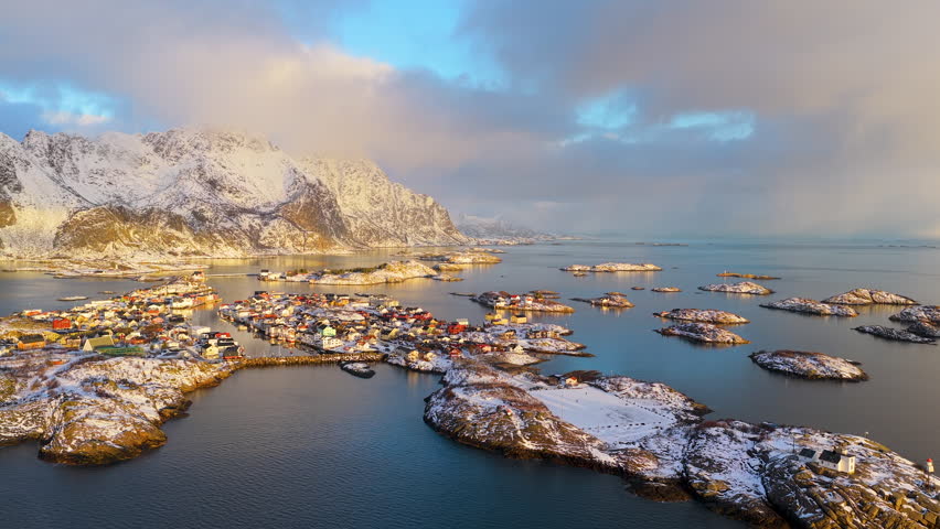 Cinematic aerial perspective of the iconic Henningsvaer fishing village surrounded by snowcapped mountains and arctic sea during golden hour in Lofoten, Norway.