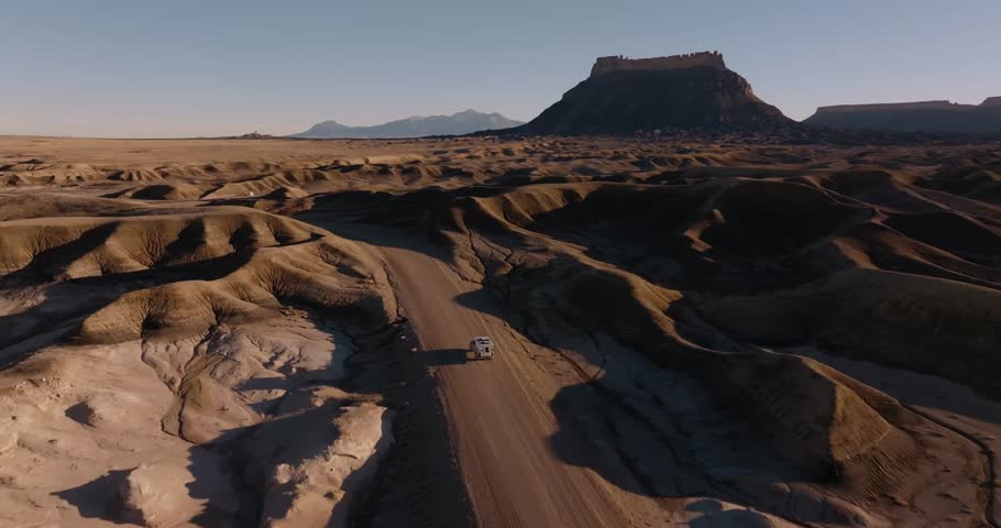 White camper van driving dirt road through eroded desert badlands near Factory Butte Utah United States, aerial rearview tracking follow