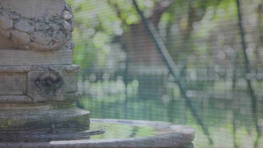 Fountain anchoring left, wire cube appearing above basin, woman walking through, AR showing design. Stone, column, outdoor, park, water, overlay, pedestrian