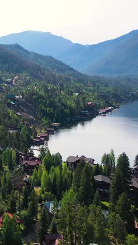 Portrait drone shot over a lakeside mountain neighborhood featuring wooded hillsides, scattered homes, reflective water, and rugged ridgelines surrounding a peaceful alpine setting