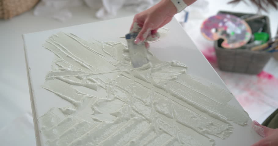 Mature woman reaching bucket spreading paste on canvas with palette knife making texture in studio. Artist, adult, tabletop, gesso, overhead, diagonal, layering