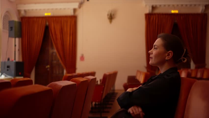 Solitary woman watching a distant stage performance in an empty theater, seated among rows of red chairs under warm ambient lighting and heavy velvet curtains