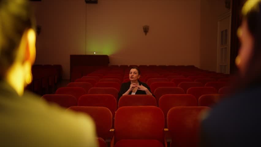 Solitary spectator in empty theater auditorium listening to presenters on stage, illuminated by warm cinematic lighting and surrounded by rows of red velvet seats
