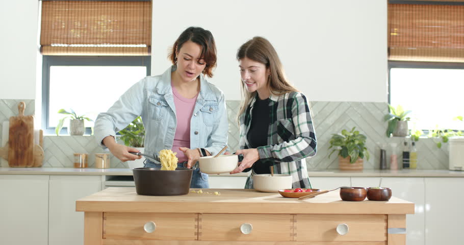 After spatula lifting pasta, female friends in denim jacket scooping into bowls in kitchen to serve. Home, cooking, duo, collaboration, plating, island, modern