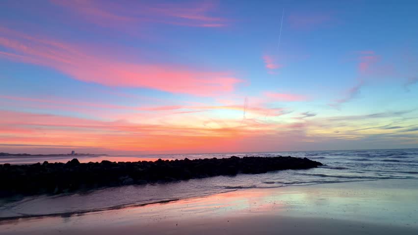 Beautiful pink and orange sunset over the ocean, with a silhouetted rock breakwater and colorful reflections on the wet sand.