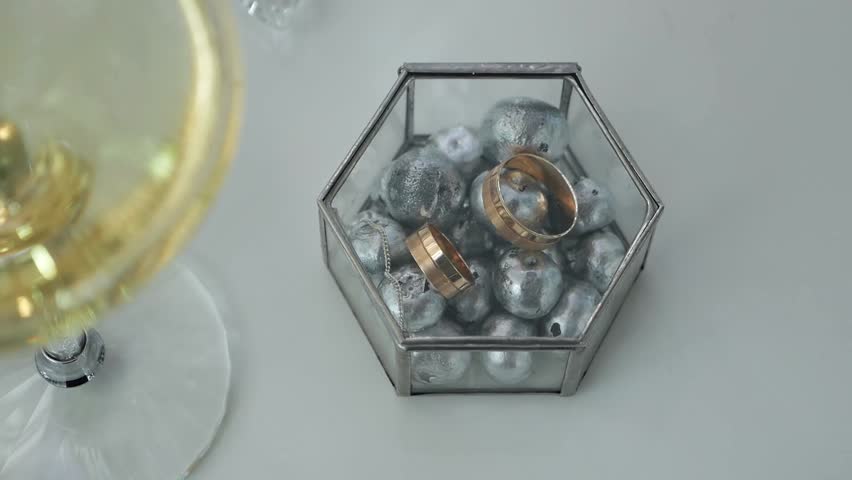 A close-up of a glass box with blueberries painted in silver with wedding rings, next to it is a glass of champagne. Wedding decor.