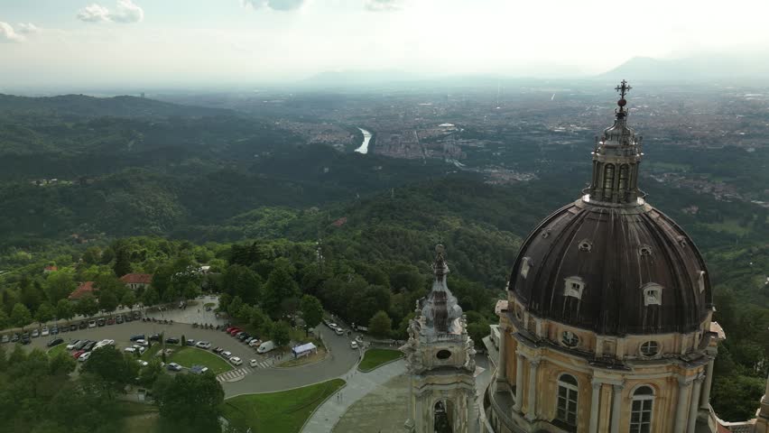Aerial drone orbit around the baroque dome of the Basilica of Superga with the city of Turin, the Po river and the plain of Piedmont visible far below in the valley.