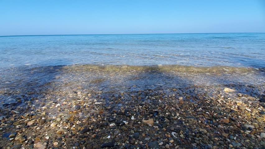 A close view of a pebble beach shoreline with small stones in the foreground and gentle waves rolling toward the coast. The clear blue sea meets a clean horizon under a bright sky, creating a calm and relaxing seascape. The texture of pebbles and transparent water adds depth and detail, perfect for concepts of nature, coastal environment, relaxation, summer atmosphere, and outdoor scenery.