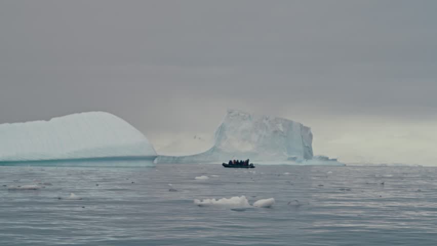 Small zodiac boat with silhouetted tourists navigating among massive Antarctic icebergs in calm grey misty polar ocean