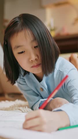A concentrated child lies on a soft carpet and draws with colored pencils on paper in a cozy home interior. The girl is engaged in creative activity, learning art skills, or spending quiet time with