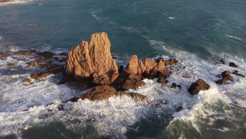 Aerial view of waves splashing on the striking rock formations of Sirens Reef in Cabo de Gata, Almeria. Warm sunset light illuminating the rugged coastline