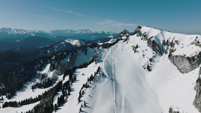 Aerial view of snow-covered mountains with sparse trees, a ski slope, and distant mountain ranges under a clear blue sky, Lenggries, Bavaria, Germany.