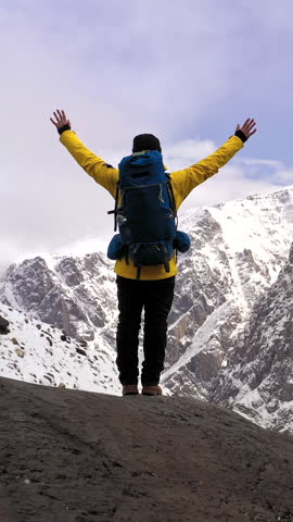 Low angle view of hiker with backpack and trekking poles standing on rocky peak with raised arms against sky. Adventurer in alpine environment during cold weather expedition. Concept of success