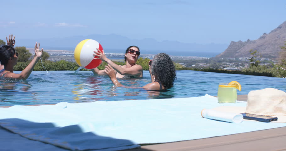 Three women passing bright beach ball, taller woman lifting, starting toss toward left for leisure. Pool, infinity, terrace, ocean, vacation, friends, sunny