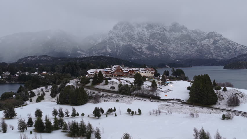 Cinematic aerial approach to Llao Llao hotel revealing snowy mountains and lake in Patagonia winter landscape.