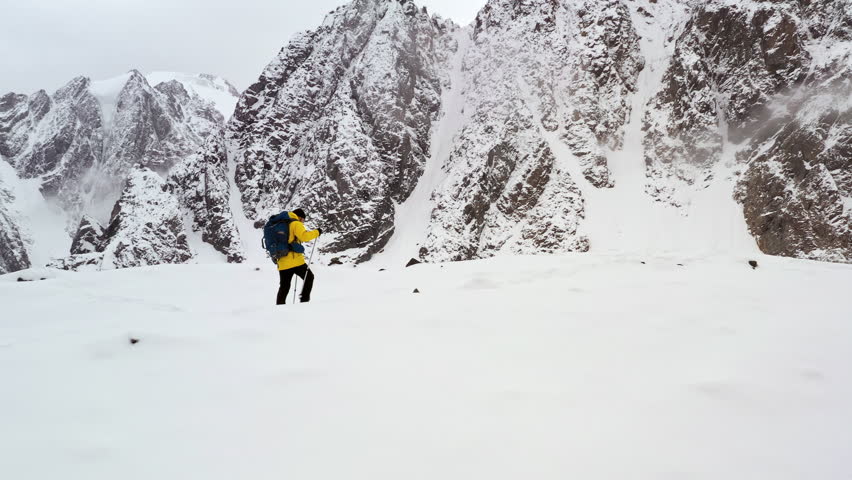 Wide shot of hiker with backpack walking across vast snowy mountain landscape with small human figure against massive cliffs. Adventurer moving through alpine terrain during cold weather expedition