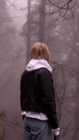 Young blonde woman in leather jacket and hoodie glancing back in moody foggy pine forest. Cinematic fashion lifestyle portrait with atmospheric grey mist and bare branches background.
