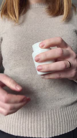 A close-up of a woman in a beige sweater pouring a single white pill or capsule from a plastic bottle into her palm to take her daily medication or vitamins.