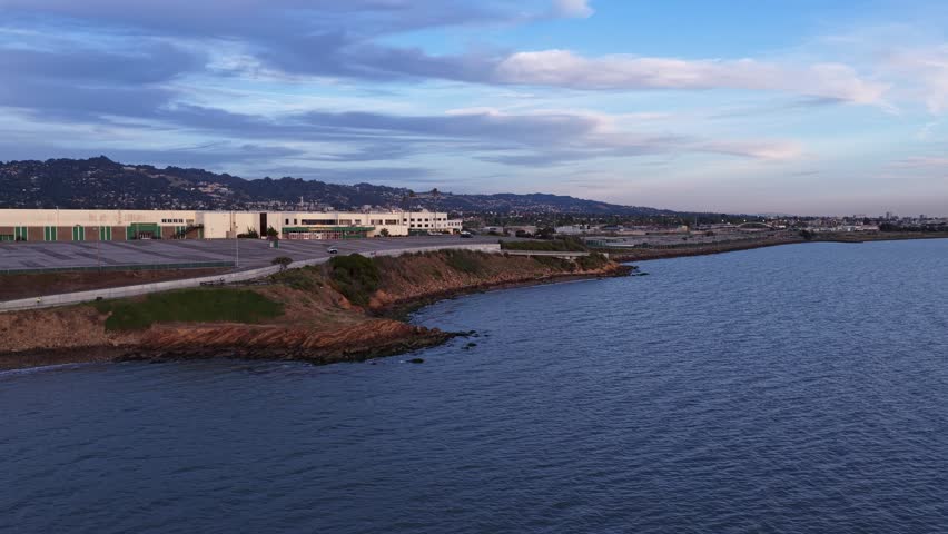 Aerial drone view moving forward over the san francisco bay towards the albany bulb, with berkeley hills in the background under a cloudy sky during a beautiful california sunset