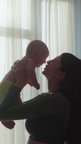 Mother joyfully lifts her baby towards the sky, creating a silhouette against bright window curtains, capturing a tender family moment