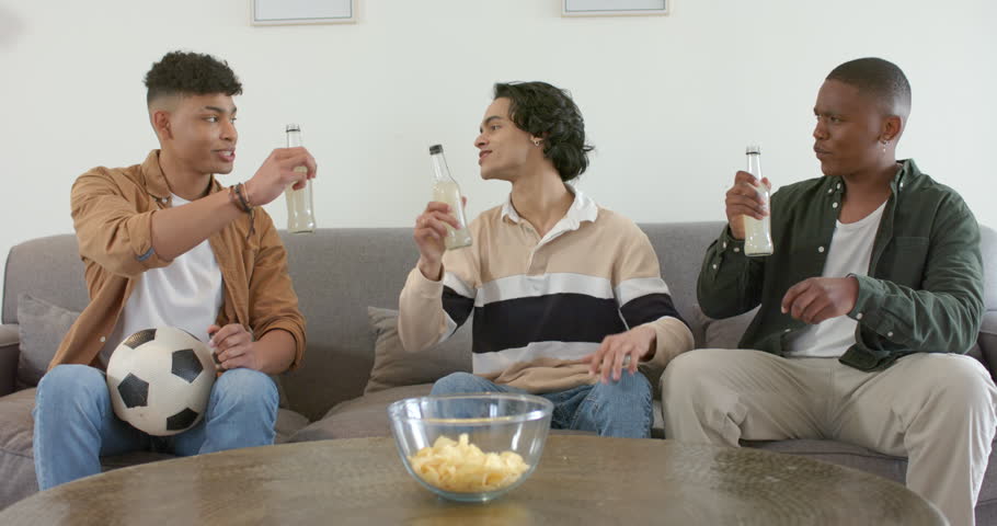 Diverse male friends holding glass bottles on sofa, clinking and opening bottle to celebrate. Celebration, camaraderie, relaxation, socializing, leisure, friendship, casual