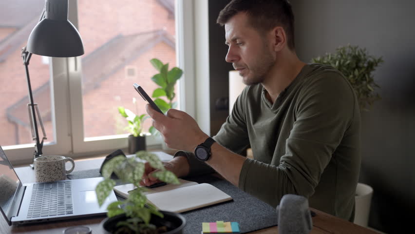 Man sitting at home desk and speaking to smartphone near laptop and notebook at home interior. Concept of voice assistant, remote work, digital routine and everyday AI technology.
