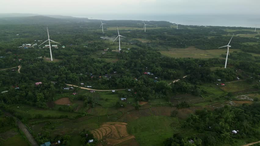 Rising aerial view of wind turbines across lush green fields with distant coastal landscape in Guimaras, Philippines during daytime.