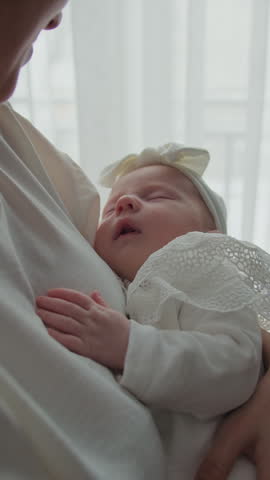 Gentle mother cradles her peacefully sleeping infant daughter, bathed in soft natural light near a window