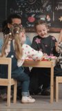 Vertical full shot of joyful Caucasian girl sitting around table with young biracial male teacher and her peers in modern kindergarten classroom and laughing during art activity - Powered by Shutterstock - Get 15% off with code: PIKWIZARD15