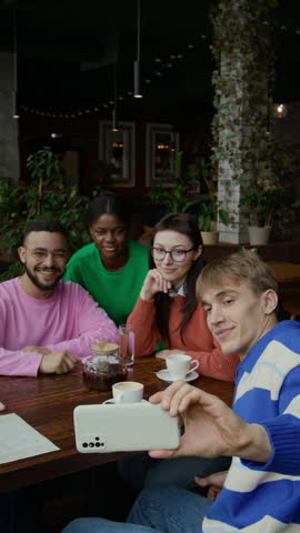 Vertical shot of four smiling young friends sitting around wooden table in cozy cafe, posing for selfie taken by gen Z man in blue and white striped shirt in foreground