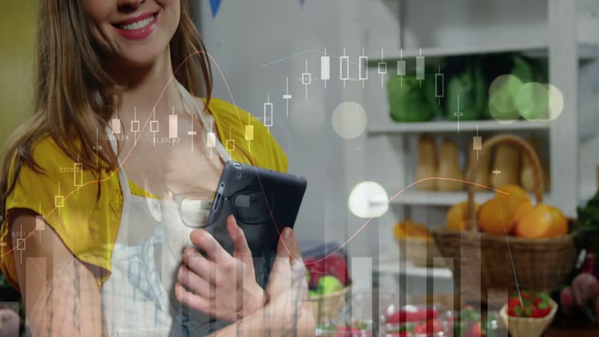 Shop worker holding tablet and starting financial charts overlaying produce while tracking sales. Market, vendor, greens, fruit, bunting, device, screen