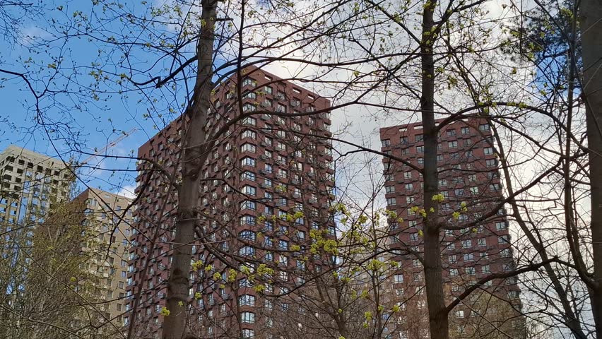 Modern high-rise residential buildings with red and beige facades seen through budding spring tree branches under bright blue sky. Urban springtime landscape. City life integrated with nature