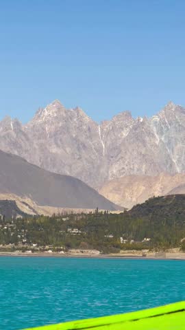 The video is from a boat sailing on Attabad Lake, Hunza, Pakistan. The backdrop features snow-capped mountains, poplar trees with yellow leaves, and the sparkling blue water on a sunny day.