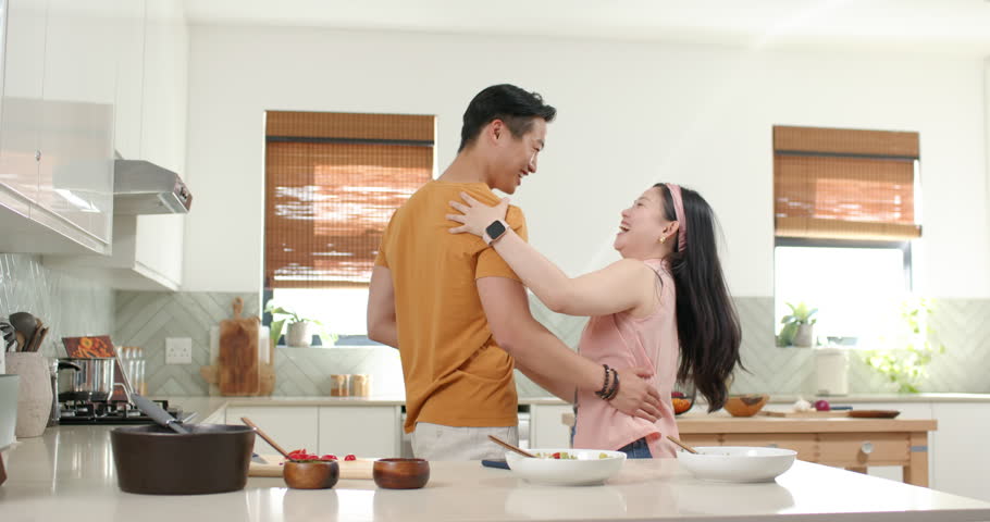 Asian couple sharing hug, swaying, and turning at kitchen island while preparing meal with bowls. Romance, affection, dance, movement, cooking, home, bright