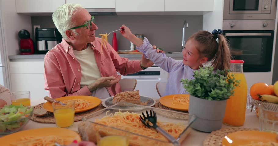 Playful little granddaughter feeding her senior grandfather spaghetti with a fork during a happy family lunch at home. Loving relationship between generations enjoying a meal together in the kitchen