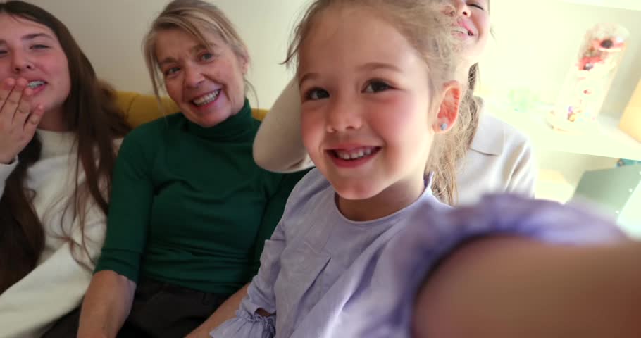 Little girl holding a camera during a video call with her family, including grandparents, mother, and aunt, all smiling and waving hello from the living room couch, capturing a joyful moment
