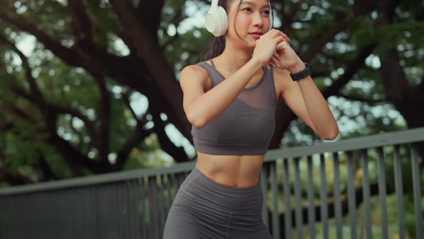Active young Asian woman in sportswear wearing white headphones and smartwatch performing squats at an outdoor park during golden hour. Concept of modern fitness, healthy lifestyle, wellness, and technology-integrated morning workout routine.