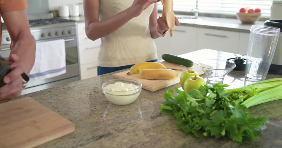 Couple reaching for blender and powder container, preparing smoothie at kitchen island. Duo, fruit, prep, slicing, peeling, yogurt, avocado