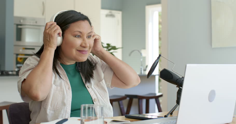 Mature Asian woman putting on white headset, adjusting mic, leaning toward laptop, starting call. Headphones, microphone, creator, podcast, remote, workspace, kitchen